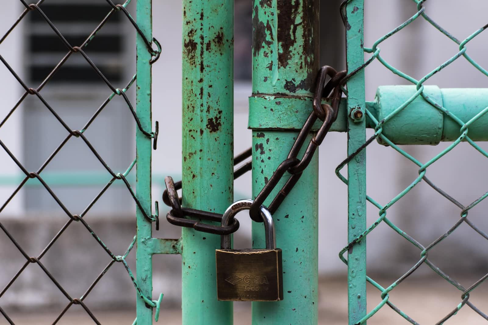 A rusty chain and brass padlock secure a green metal gate, which is part of a wire fence—much like how proper Spring transaction handling ensures security in a system. The background is blurred, showing a building.