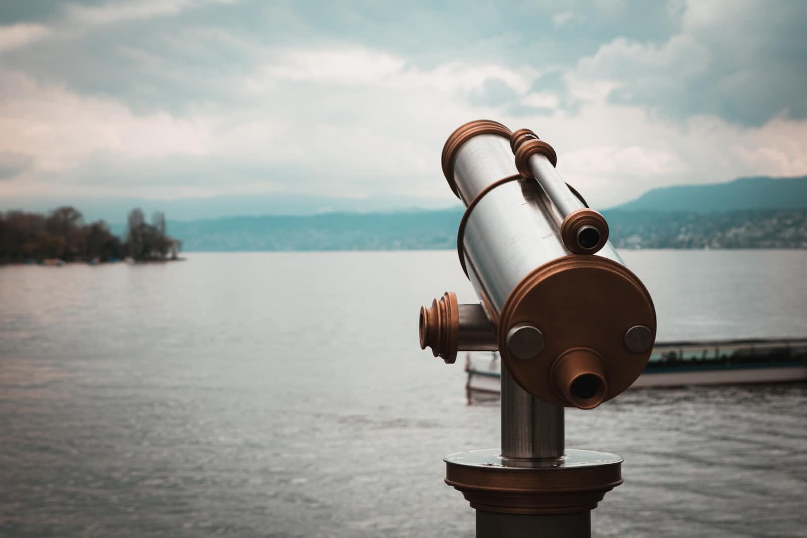 A coin-operated viewing telescope faces a calm lake under a cloudy sky, reminiscent of the way Kubernetes orchestrates resources, with distant shoreline and hills in the background.