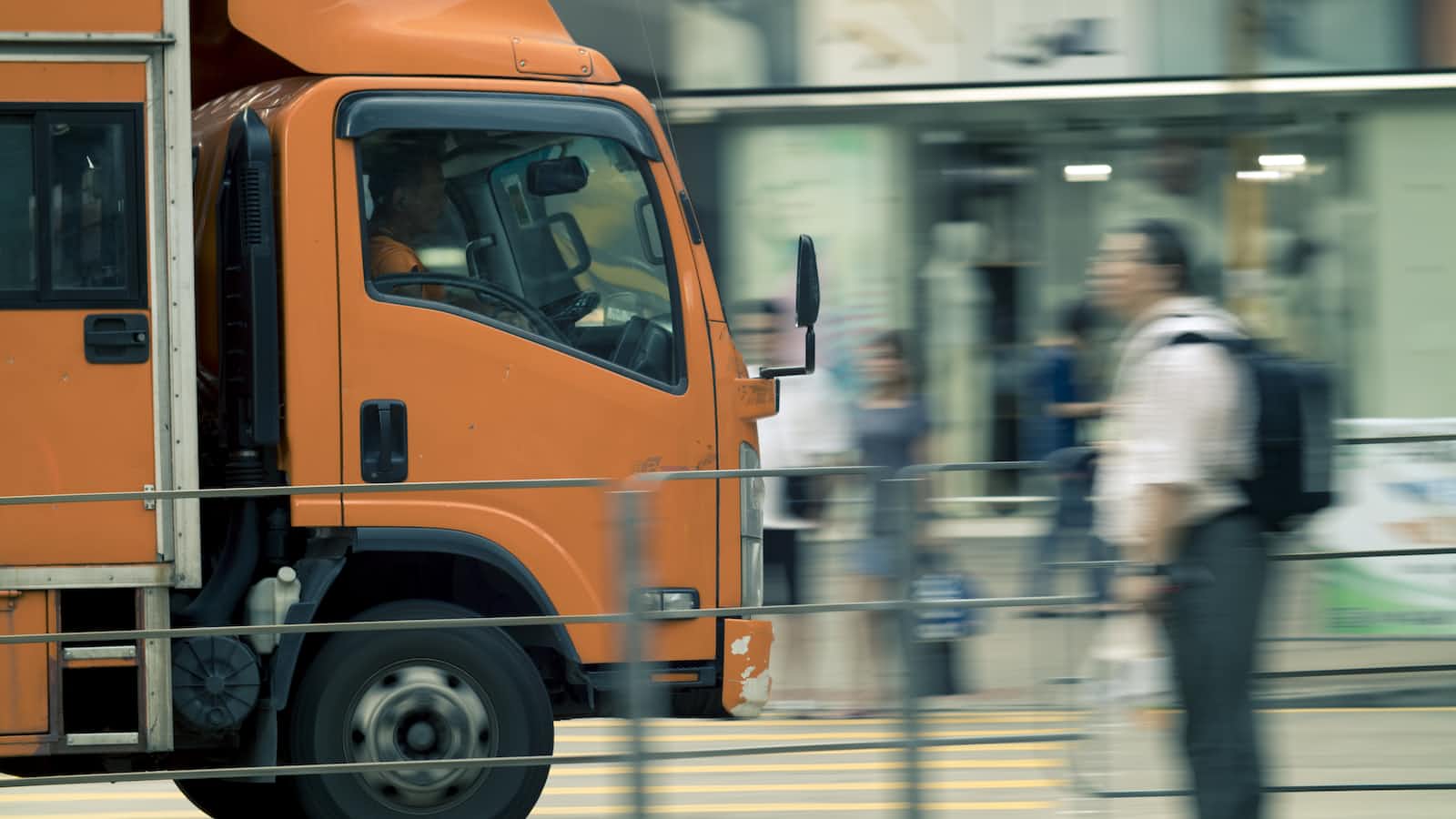 An orange delivery truck drives past a blurred pedestrian on a city street, with shop windows visible in the background, evoking the dynamic pace of cloud migration in today’s Azure-powered cities.