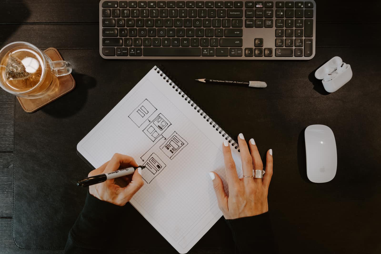 A person sketches website wireframes on grid paper at a dark desk, with a keyboard, mouse, pen, drink, and wireless earbuds nearby—gathering architectural insights to guide the next steps in web application development.
