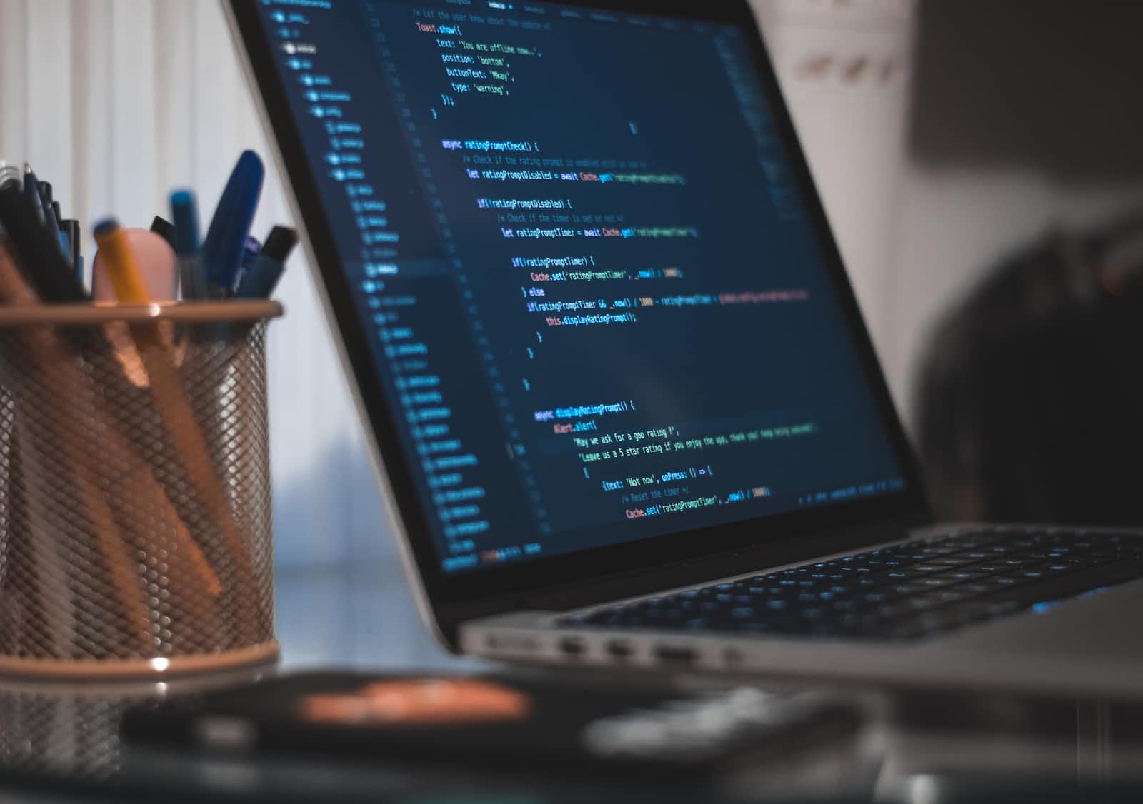 A laptop displays Vue.js code on its screen, while a metal mesh cup holding pens and pencils sits nearby on the desk—a perfect setup for rapid development. The background is softly blurred.