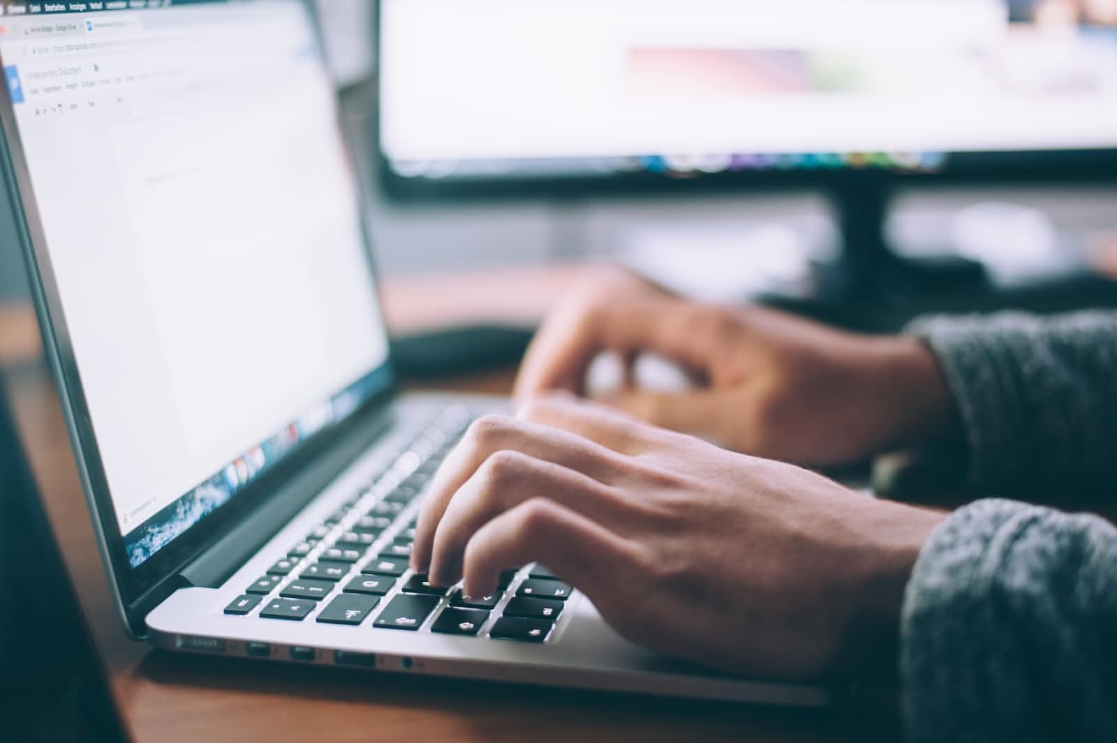 Close-up of a person typing on a laptop keyboard, with another computer monitor in the background. The scene suggests working, studying, or browsing on a desk—perhaps engaged in rapid development using tools like React-Admin or Fastify.