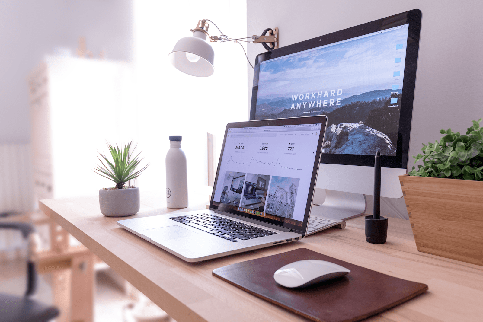 A tidy desk with a laptop, desktop monitor, mouse, mouse pad, water bottle, potted plant, lamp, and a pen in a holder. The monitor displays a scenic wallpaper alongside a Microsoft Power BI dashboard and the words WORK HARD ANYWHERE.