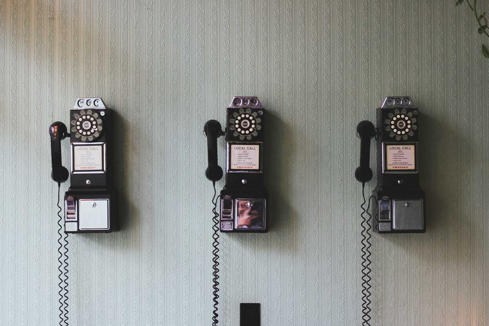 Three vintage black rotary payphones are mounted on a light green patterned wall, each with a coiled cord and a metal coin slot below the rotary dial, evoking classic interaction modes from an earlier era.