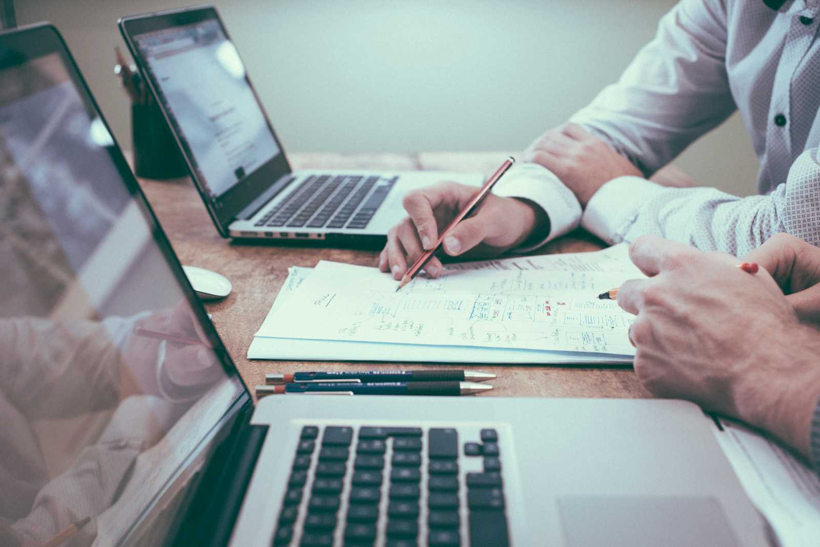 Two people review documents and take notes at a desk with two open laptops, pencils, and papers, suggesting a collaborative work or study session focused on Requirements Engineering in an office setting.