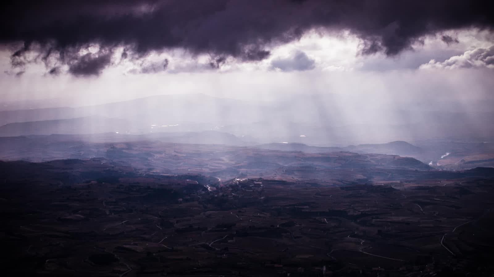 Dark clouds cover the sky as sun rays break through, casting light over a vast, hilly landscape below. Much like the refined interface of Angular Material 19, the scene appears dramatic and moody, with soft illumination highlighting the terrain’s natural contours.