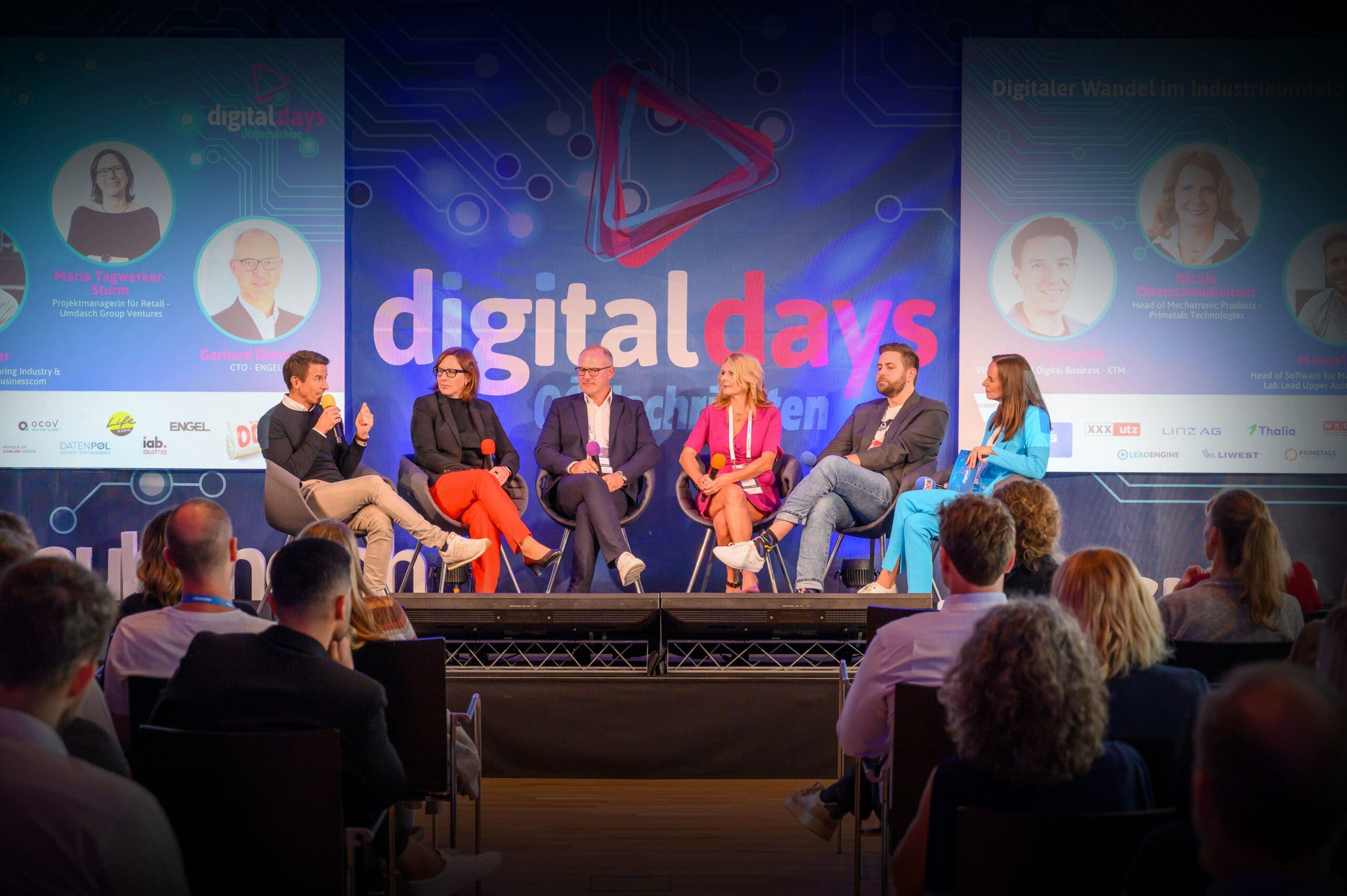 A panel of six people sits on stage at the OÖN Digital Days conference, facing an audience while a speaker addresses them with a microphone.